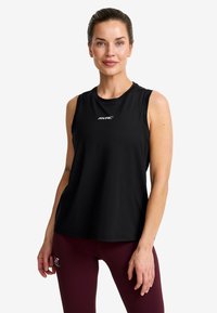 Woman with pulled-back hair wearing a black sleeveless top and burgundy leggings, standing and facing forward against a white background.