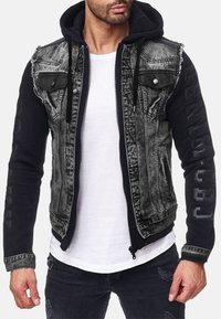 Giacca in denim nero e tessuto con cappuccio, caratterizzata da dettagli distressed, tasche frontali e ricami sulle maniche.