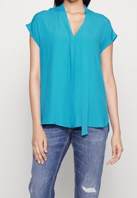 Blusa azzurra a maniche corte con scollo a V e dettaglio annodato davanti. Realizzata in tessuto leggero, presenta una vestibilità morbida e una superficie liscia.