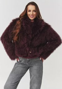 Burgundowa kurtka z sztucznego futra z przytulnym kołnierzem, w oversize'owym fasonie, z zapięciem na guziki. Noszona z szarymi dżinsami, o miękkiej, teksturowanej powierzchni.