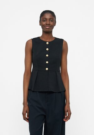 Femme portant un haut noir sans manches avec cinq boutons dorés sur le devant et un pantalon foncé, debout devant un fond blanc uni.