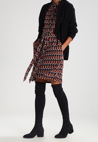 Robe manches longues avec un motif géométrique rouge, noir et crème, ceinturée à la taille, associée à des bottes plates noires montantes jusqu'aux genoux et un cardigan noir.