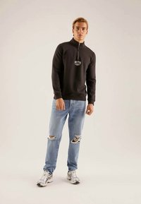 Melna džemperis ar rāvējslēdzēju kakla daļā un uzrakstu "BOSTON", kopā ar gaiši zilām saplēstām džinsu biksēm. Modelis valkā baltus un pelēkus sporta apavus.