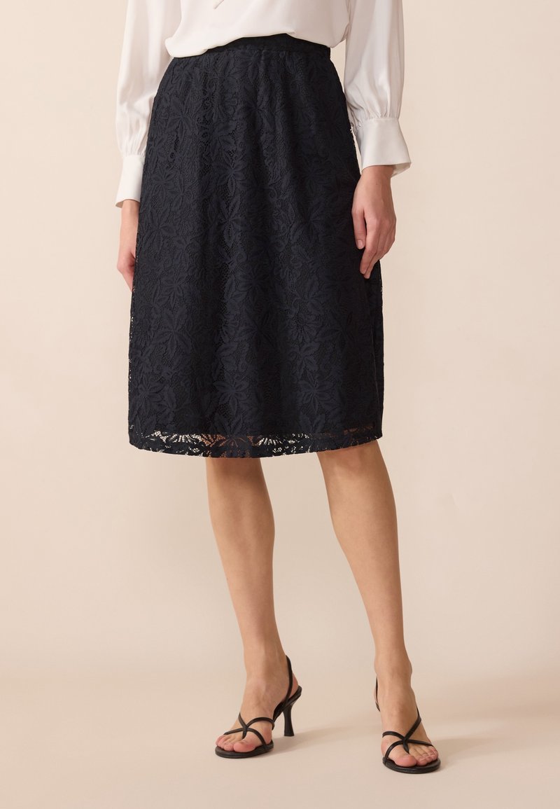 Navy lace skirt with floral pattern, elastic waistband, and scalloped hem, paired with black strappy heels and a white blouse.