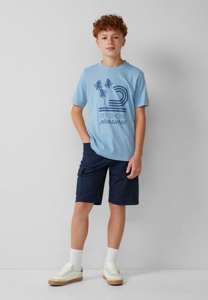 T-shirt en coton bleu clair avec un graphique de palmier et le texte "OFFSHORE pleasures", accompagné de shorts cargo navy et de baskets blanches.