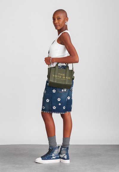 Olijfgroene leren tote bag met gouden letters, gecombineerd met een spijkerrok met witte bloemengebras en blauwe distressed high-top sneakers.