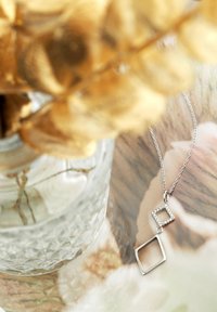 Silverhalsband med två geometriska hängen: en fyrkant med klara stenar och en öppen fyrkant. Mot en suddig blomsterbakgrund.