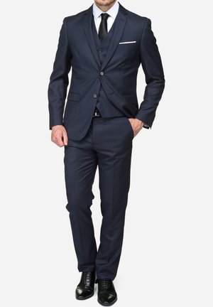 Homme portant un costume trois pièces bleu marine avec une cravate noire, une chemise blanche, des chaussures noires et un mouchoir blanc dans la poche, main dans la poche.
