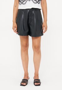 Pantalones cortos de cuero negro con una textura suave y un diseño a medida. Presentan pliegues en la parte delantera y un ajuste ceñido. Se combinan con sandalias negras de tiras.