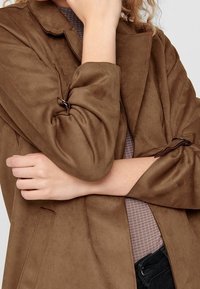 Veste marron en suédine synthétique avec un large col et des manches retroussées, dotée d'un détail à nouer et d'une texture lisse. Portée sur un haut à carreaux.