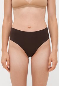 Woman wearing high-waisted dark brown seamless underwear and a beige bra, standing with arms relaxed at sides against white background.