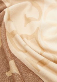 Sciarpa intrecciata con morbidi colori beige e marrone chiaro, caratterizzata da un sottile motivo geometrico, che mostra una texture liscia e un tessuto che si drappeggia.