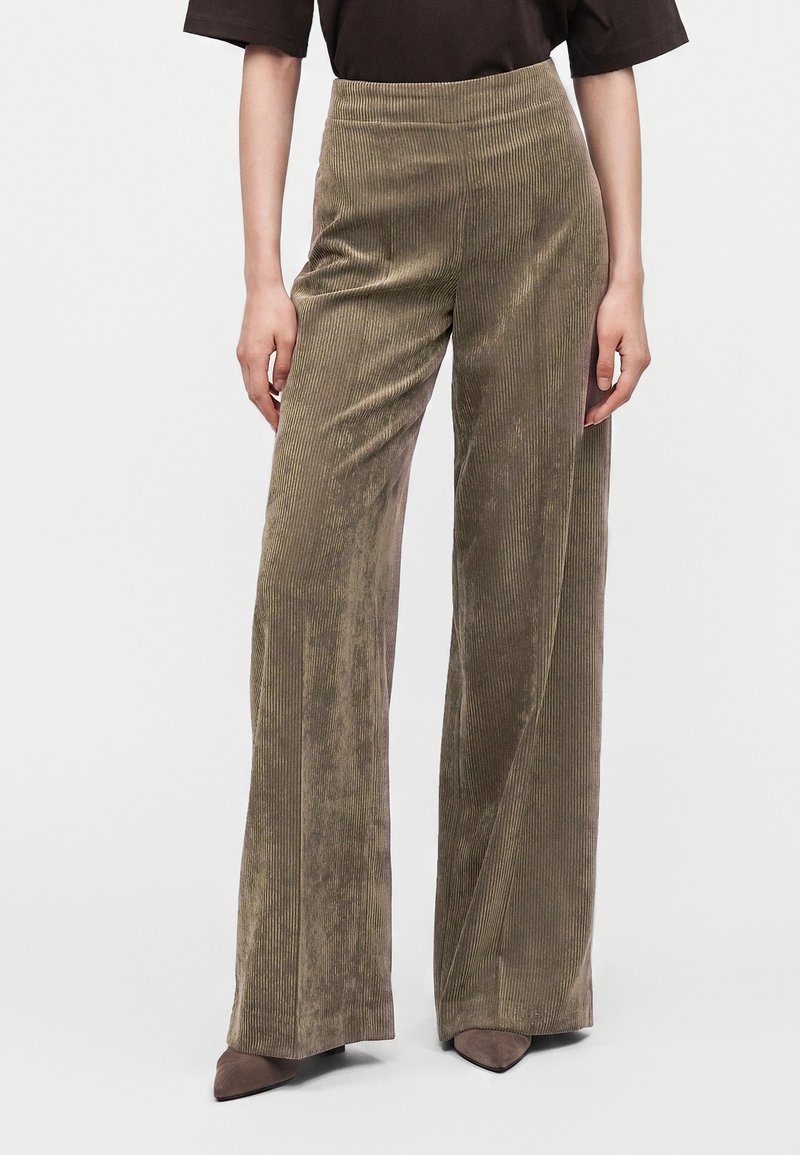 Femme portant un pantalon en velours côtelé taille haute à jambes larges, de couleur vert olive avec des rainures verticales, assorti à des chaussures marron à bout pointu.