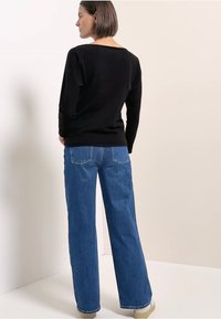 Czarna bluzka z długim rękawem i wycięciem na plecach, zestawiona z szerokimi, niebieskimi dżinsami. Wyraźne przeszycia na kieszeniach i dole. Neutralne tło.