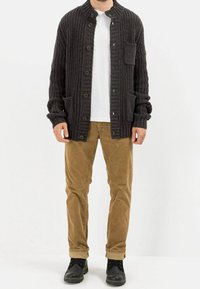 Cardigan en tricot gris foncé avec col montant, boutons sur le devant, deux poches, porté sur une chemise blanche, associé à un pantalon en velours côtelé beige et des bottes noires.