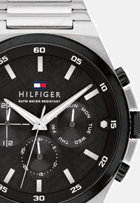 Montre en acier inoxydable argenté avec un cadran noir texturé, des index horaires blancs et des sous-cadrans pour le jour et la date. Présente une étanchéité de 5ATM.