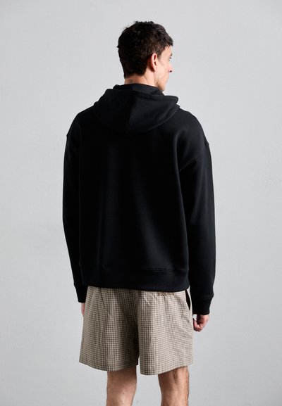 Schwarzer Kapuzenpullover mit lockerer Passform, ausgestattet mit einer Kängurutasche und gerippten Bündchen. Kombiniert mit braunen Karokurzshorts.