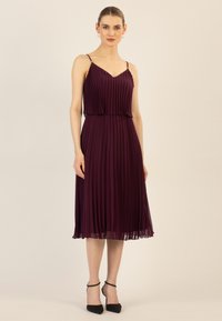 Bordeauxfarbenes plissee Kleid mit einem V-Ausschnitt, dünnen Trägern und einem ausgestellten Saum. Kombiniert mit schwarzen Absätzen mit Knöchelriemen, was einen kontrastierenden Look erzeugt.