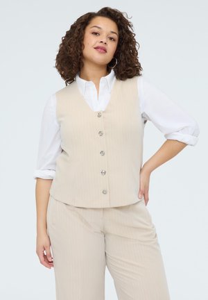 Kiharatukkainen nainen, jolla on päällä beige nappikiinnitteinen liivi ja siihen sopivat housut valkoisen, hihoilta käärityn paidan päällä, seisoo yhdellä kädellä lantiolla.