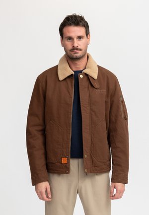 Man draagt een bruine jas met een beige shearling kraag, navy trui en beige broek, staand tegen een effen lichte achtergrond.