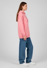 Felpa con cappuccio rosa chiaro, dotata di tasca frontale, abbinata a jeans a gamba ampia blu e sneakers bianche, su sfondo grigio semplice.
