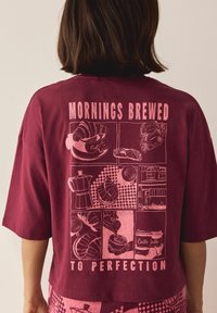 Bordo īsām piedurknēm krekls ar rozā grafikas dizainu, kas ietver ēdienu un kafijas ilustrācijas, teksts lasīts "RĪTI RECEPTES, KAS IEVĒROTAS LIEKAM."