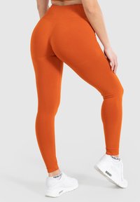 Leggings a vita alta e aderenti di un arancione vibrante, con una texture liscia e una cintura elastica, abbinati a scarpe da ginnastica bianche.