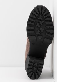 Semelle en caoutchouc noire avec des sculptures profondes et des motifs triangulaires sur une botte en daim taupe reposant verticalement contre une surface blanche.