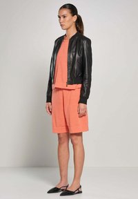 Giacca bomber nera in pelle su una maglietta corta corallo e pantaloni corti abbinati, completata da ballerine nere a punta. Design semplice e moderno.