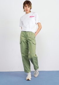 T-shirt blanche à manches courtes avec logo rose, associé à un pantalon cargo vert olive avec poches latérales et une coupe ample, porté avec des baskets blanches.