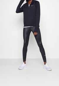Svart zip-up sporttop, slim-fit blanka svarta leggings och vita sneakers. Toppen har en krage; leggings har en subtil lyster och logotyp.