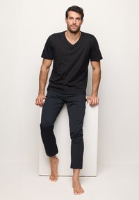 Zwarte v-hals t-shirt gecombineerd met donkere slim-fit broek. Glad oppervlak, minimalistisch design. Model staat blootsvoets tegen een neutrale achtergrond.