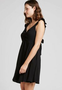 Robe noire avec un corsage ajusté, des bretelles volantes et une jupe évasée. Confectionnée en tissu léger, elle présente un décolleté en V et des fronces à l'avant.