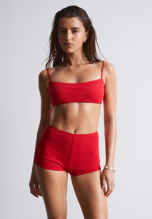 Femme aux cheveux mouillés portant un crop top côtelé rouge et un short taille haute assorti, debout devant un fond clair uni.
