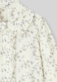 Blouse florale en tissu ivoire transparent, avec un col volanté, des boutons sur le devant et des motifs de fleurs grises délicates. Légère et texturée.