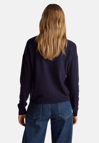 Suéter de punto azul marino con puños y dobladillo acanalados, combinado con unos vaqueros de mezclilla azul de cintura alta que cuentan con bolsillos traseros y una etiqueta verde con el logo.