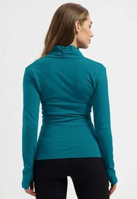 Türkisfarbena, langärmelige Sportoberteil mit hohem Kragen, taillierter Passform, glatter Textur und Raffung im Rücken. Kombiniert mit schwarzen Hosen.