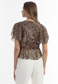 Gennemsigtig brun bluse med slangeprint, bølgede ærmer og en indsnævret talje. Kombineret med hvide bukser, der viser en peplum-silhuet.