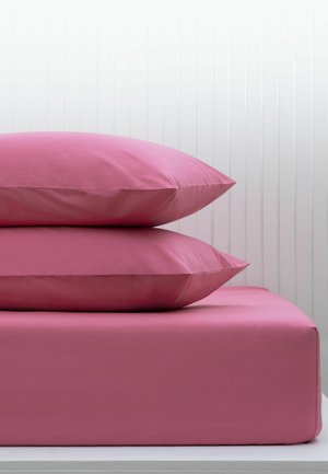 Zwei rosa Kissen, gestapelt auf einem rosa Spannbettlaken auf einem Bett mit einer weißen vertäfelten Wand im Hintergrund.