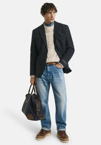 Uomo che indossa un blazer scuro, un maglione a trecce color avorio, jeans azzurri chiari e scarpe marroni, mentre tiene una borsa da weekend blu navy e marrone.
