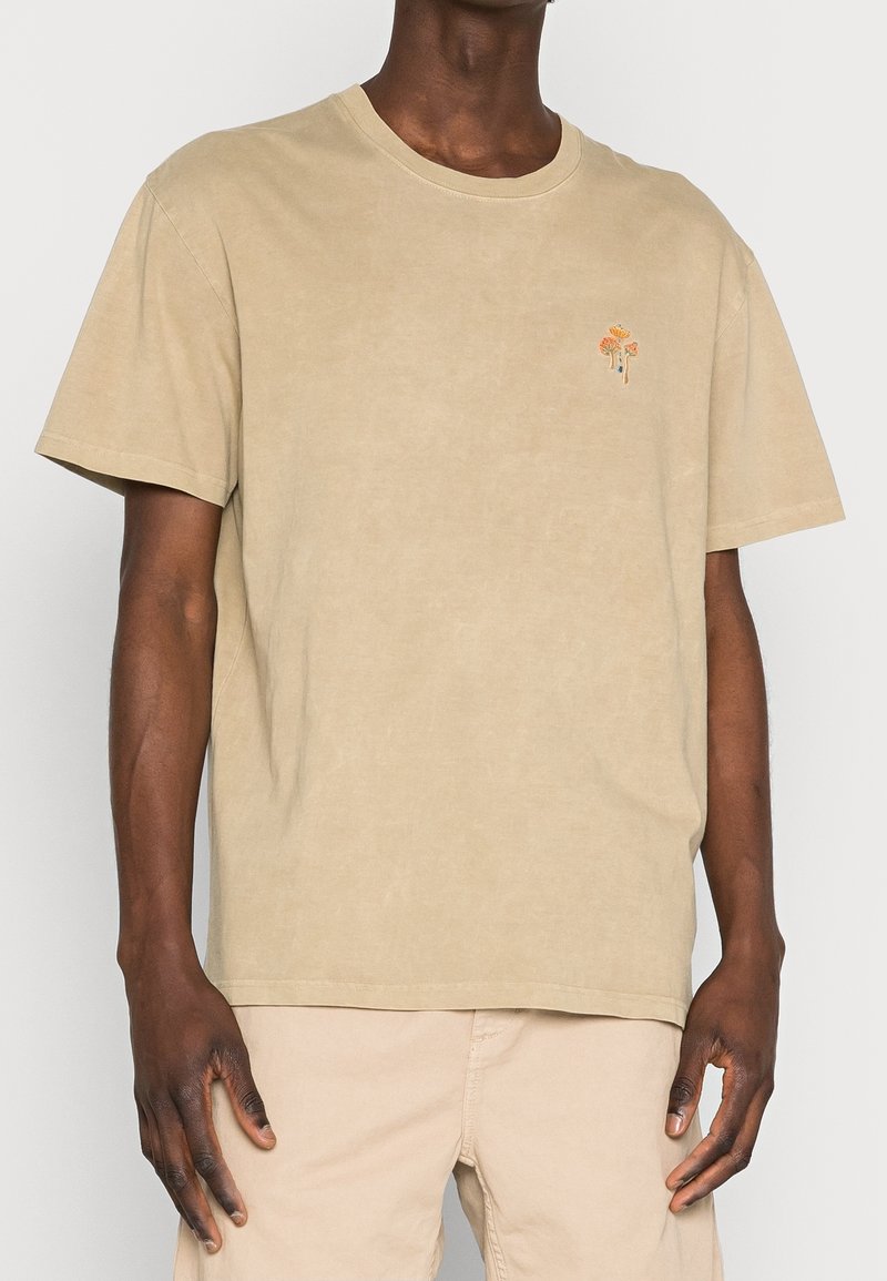 Muž v béžovém tričku s krátkými rukávy, na levé straně hrudi je malé vyšívané oranžové květinové motiv. K tomu nosí béžové kalhoty.