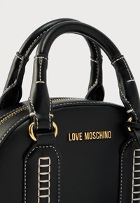 Μαύρη τσάντα από συνθετικό δέρμα με διπλές δομημένες λαβές, χρυσές λεπτομέρειες λογότυπου, λευκές ραφές και επάνω κλείσιμο με φερμουάρ.