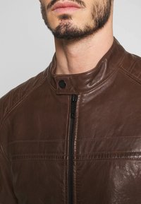 Hnědá kožená bunda s vysokým límcem, prošívaným designem ramen a předním zipem. Obsahuje černé kovové detaily a hladkou texturu.