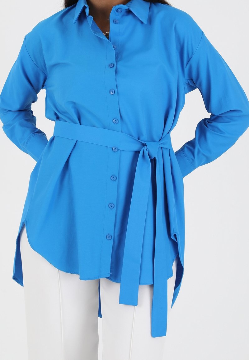 Femme portant une chemise bleue à manches longues boutonnée avec une ceinture nouée à la taille et un pantalon blanc, les mains sur les hanches.