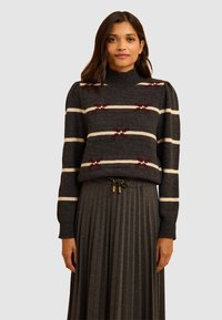 Maglione grigio a maglia con strisce bianche e bordeaux, colletto alto e maniche a sbuffo, abbinato a una gonna plissettata grigia con vita regolabile.