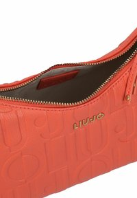 Sac à main orange en cuir texturé, présentant des motifs embossés, une fermeture éclair dorée et un accent logo. L'intérieur est doublé de tissu.