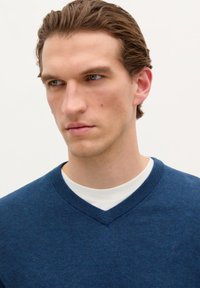Maglione in maglia di colore blu navy con scollatura a V, dotato di bordi a coste e un piccolo logo sul petto. Indossato sopra una camicia bianca.