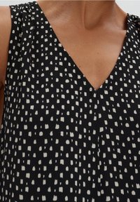 Musta kangastoppi, jossa on V-nahka-design, ja epäsäännöllisiä valkoisia pisteitä, pehmeä rakenne ja laskostetut olkaimet.