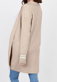 Kvinna klädd i en beige öppen lång cardigan med fickor och ljusblåa ränder på ärmarna, tillsammans med blå jeans.