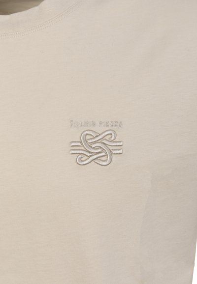 Hellbeiges Stoff mit weißem, gesticktem, ineinander verschlungenem Knoten und dem Text "Filling Pieces" über dem Design im Brustbereich.